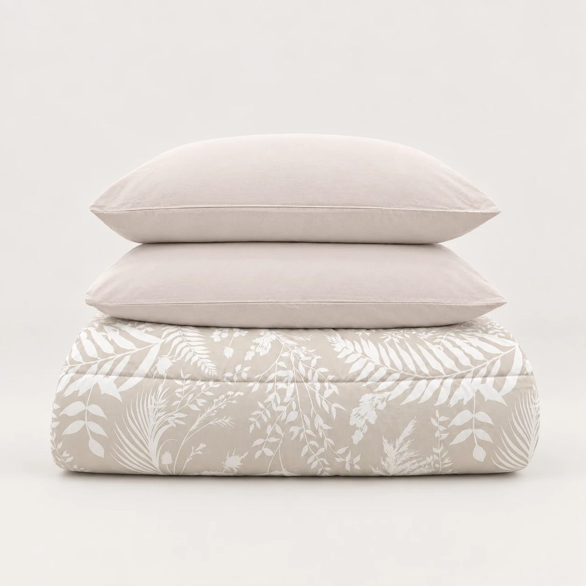 Edredón de invierno beige con estampado de hojas blancas doblado impecablemente, coronado por dos fundas de almohada a juego.